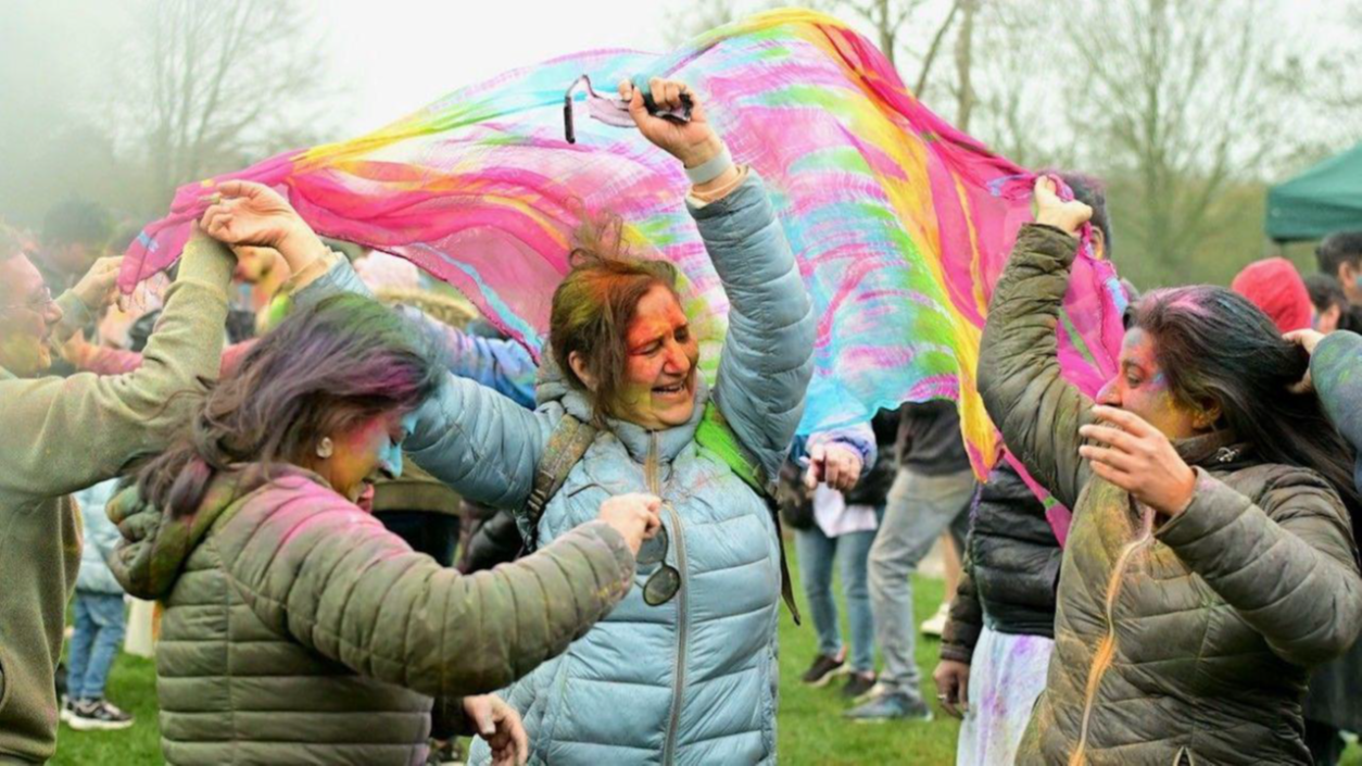People standing outdoors at a public event hold a large, brightly coloured fabric sheet above their heads. Several people are gathered close together underneath and around the fabric, wearing coats. The fabric is multicoloured with pink, green and yellow tones, and stretches across the group, creating a canopy effect. Other people are visible in the background on a grassy area with trees and a tent.