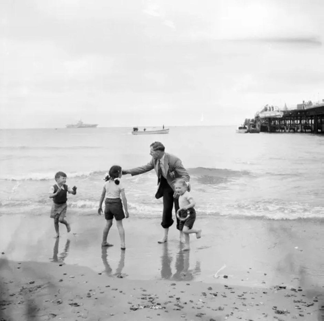 Black and white photo of an adult and three children at the water’s edge on a beach, with a pier and boats in the background.